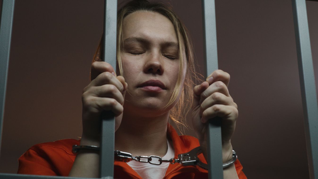Female,Prisoner,In,Orange,Uniform,Holds,Metal,Bars,,Stands,In