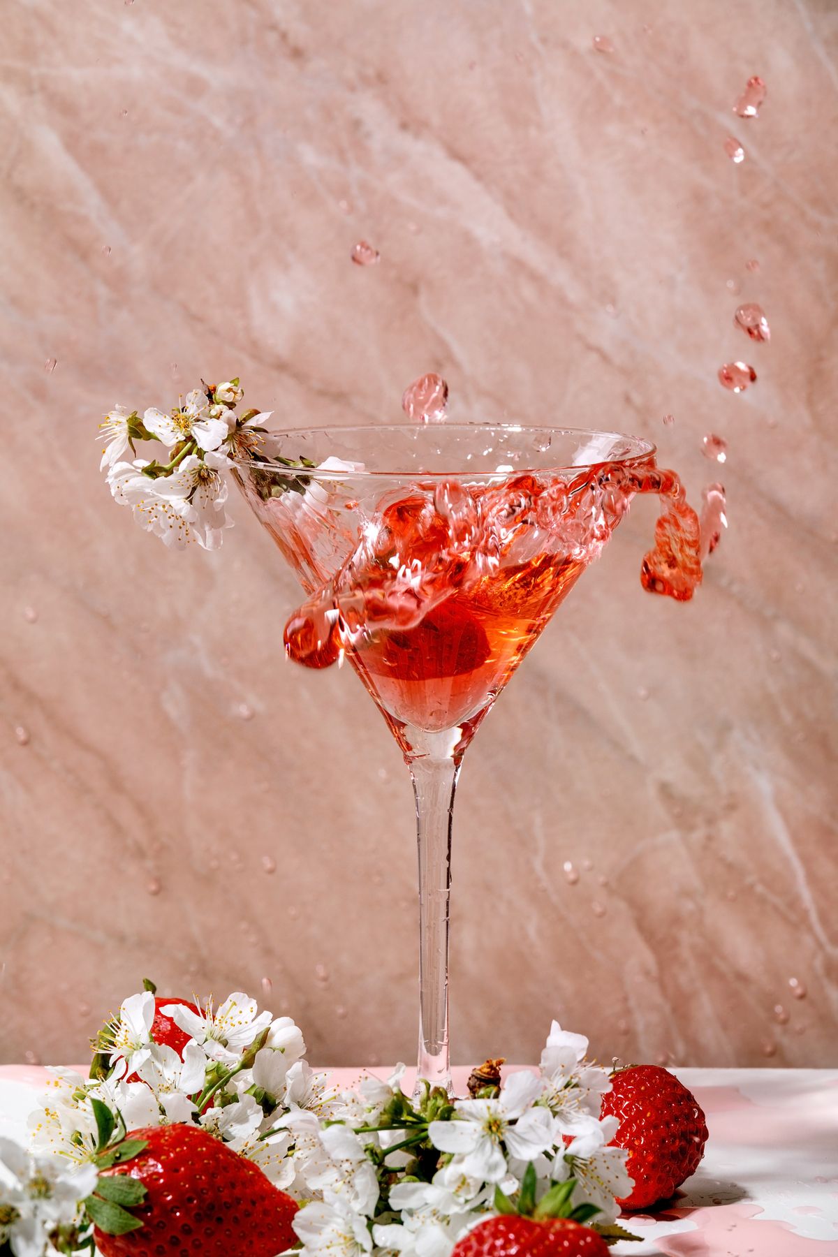 Splash of strawberry alcoholic or non-alcohol cocktail in martini glass decorated with blooming branches of cherry tree over white and pink texture background