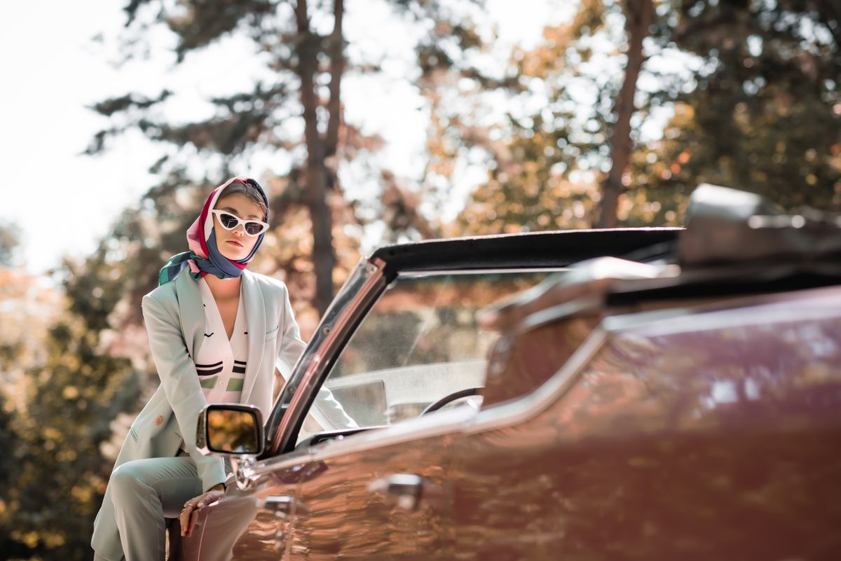 Stylish,Woman,Sitting,On,Car,On,Blurred,Foreground,Outdoors