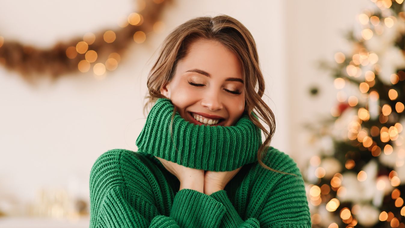 Portrait of a funny emotional cheerful young woman with horns on her head in a green cozy warm sweater laughing having fun holding and eating a lollipop and ginger cookies on Christmas day on the background of a decorated Christmas tree at home in winter