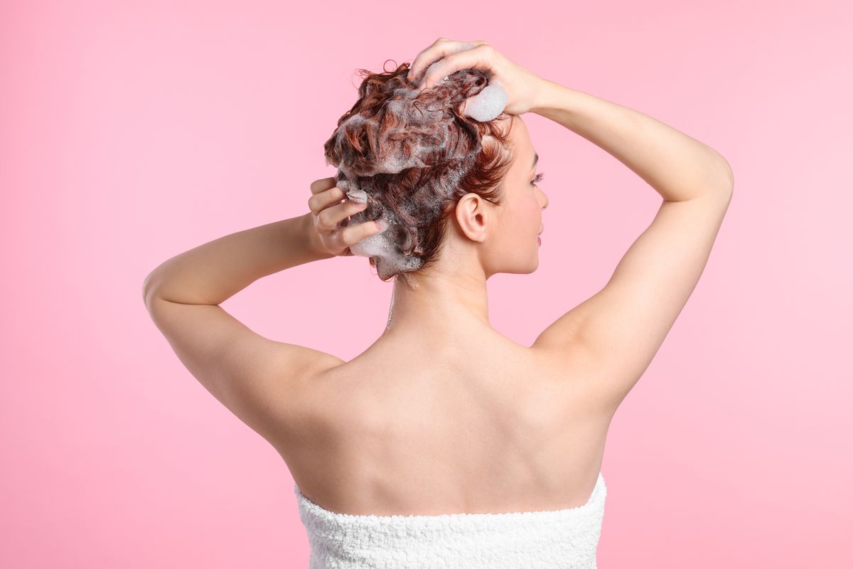 Young woman washing her hair with shampoo on pink background, back view. hajmosás
