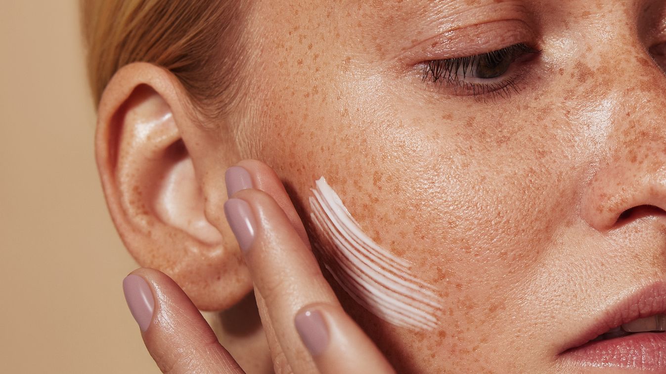 Close up highly detailed shot of a young woman applying cream on her freckled skin. Cropped shot of woman applying moisturizer on cheek with fingers.