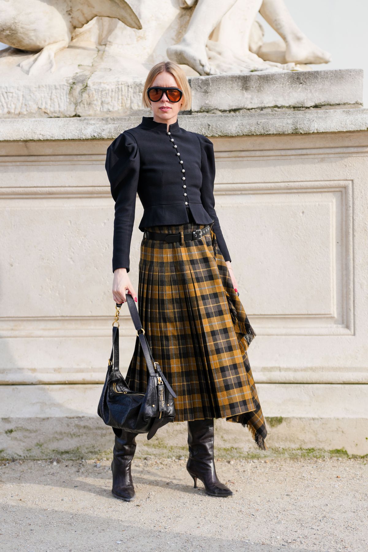 PARIS, FRANCE - MARCH 04: A guest wears black frame dark orange lenses sunglasses, black tailored corset puffy long sleeve Dior blazer jacket, shiny black Dior leather bag, shiny black leather belt, dark yellow plaid pattern/print scarf midi skirt, shiny black pointed toe slouchy heels leather shoes, outside Christian Dior, during the Paris Fashion week Women's Fall/Winter 2025-2026 on March 4, 2025 in Paris, France. (Photo by Edward Berthelot/Getty Images)