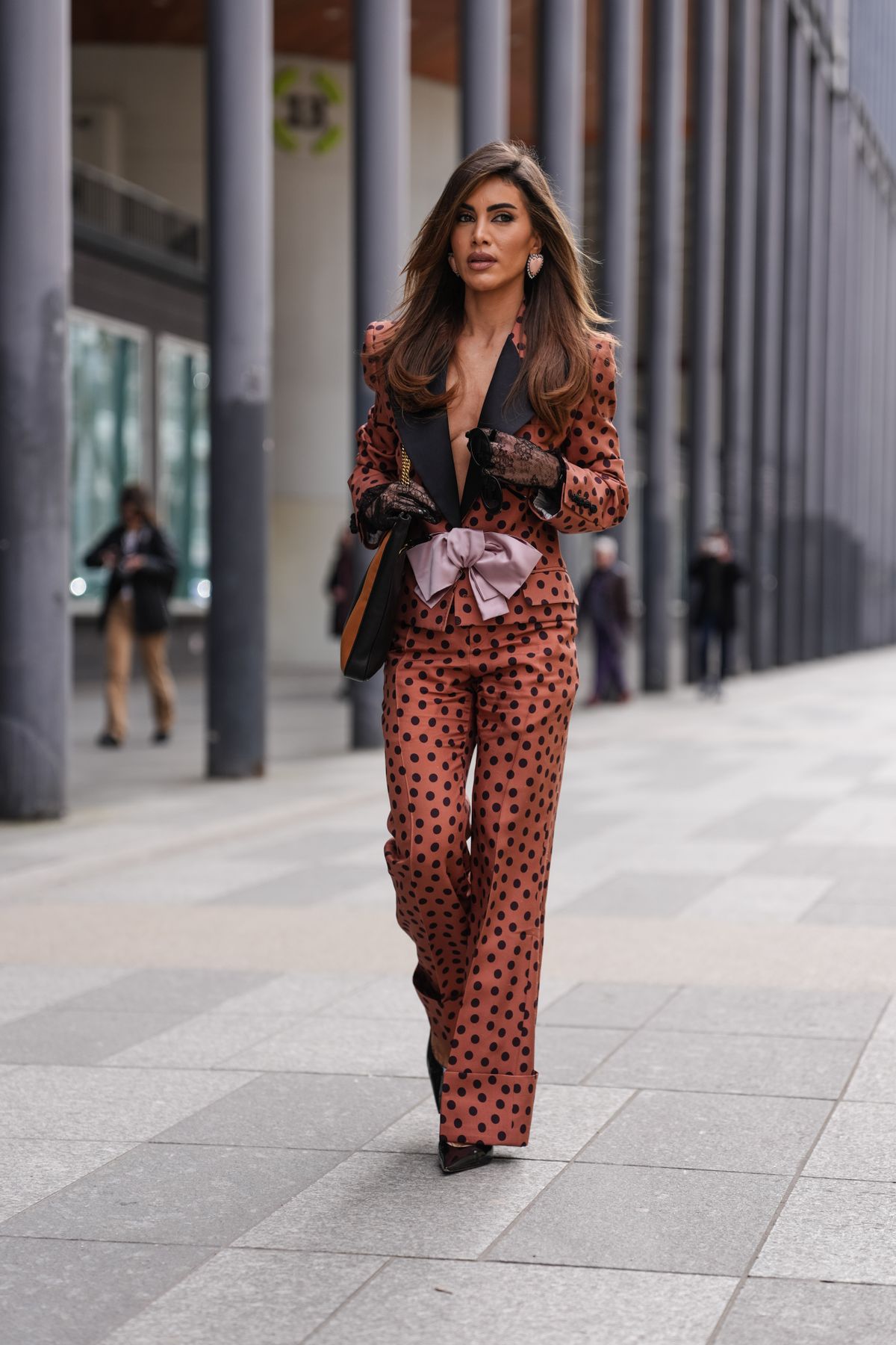 PARIS, FRANCE - MARCH 09: Camila Coelho wears light pink heart shaped earrings, dark brown black polka dotted light pink bow Valentino blazer jacket, black dark brown pattern Valentino leather bag, black sheer mesh lace gloves, dark brown black polka dotted light pink bow Valentino relaxed trouser pants, black pointed toe heels leather shoes, outside Valentino, during the Paris Fashion week Women's Fall/Winter 2025-2026 on March 9, 2025 in Paris, France. (Photo by Edward Berthelot/Getty Images)