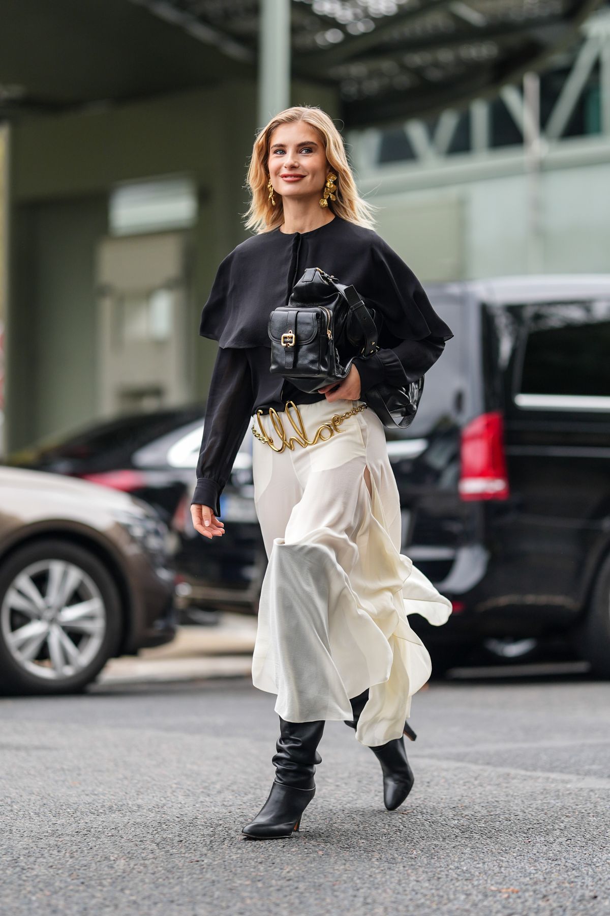 PARIS, FRANCE - SEPTEMBER 26: Xenia Adonts wears black long sleeve shirt, shiny black Chloe leather bag, gold Chloe belt, white sheer mesh flowy asymmetrical Chloe midi skirt, shiny black thigh high heeled leather boots, outside Chloe, during the Paris Fashion Week Spring/Summer 2025 on September 26, 2024 in Paris, France (Photo by Edward Berthelot/Getty Images)