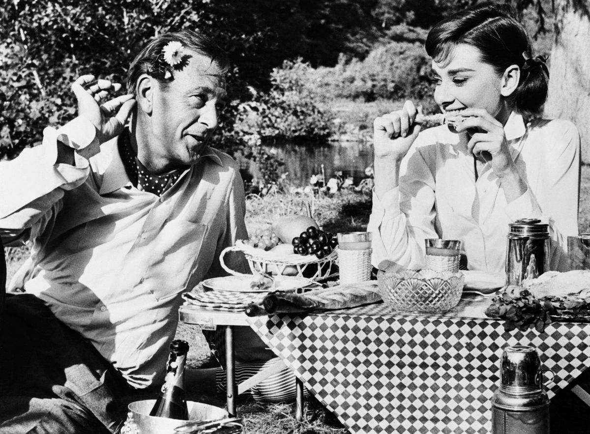 (Original Caption) 10/1/1956-Paris, France: Gary Cooper, playing the role of a philandering American, is shown wooing Audrey Hepburn at a De Luxe picnic for two in their new movie "Love in the Afternoon." Coop is trying to impress the lady by wering a daisy over his ear, but she goes calmly chewing on a drumstick. (Photo by Bettmann via Getty Images)