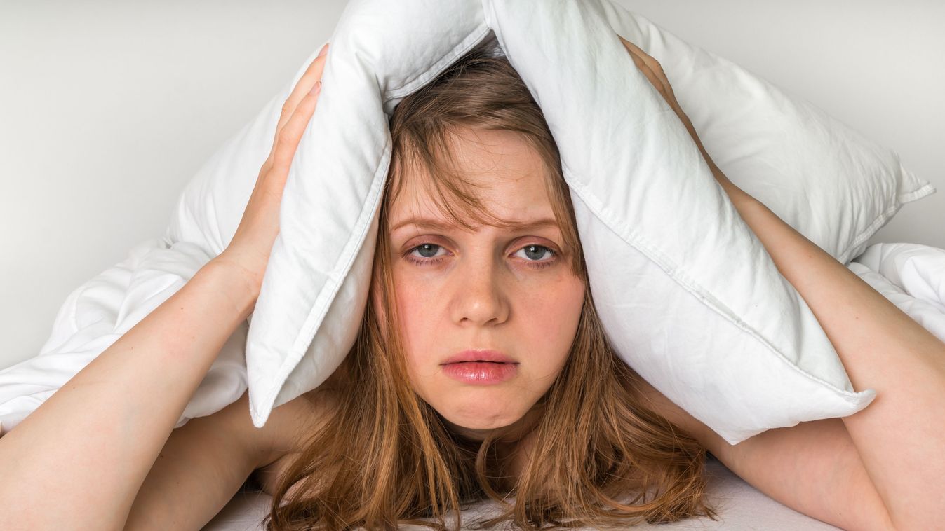 Young woman in bed covering ears with pillow because of noise