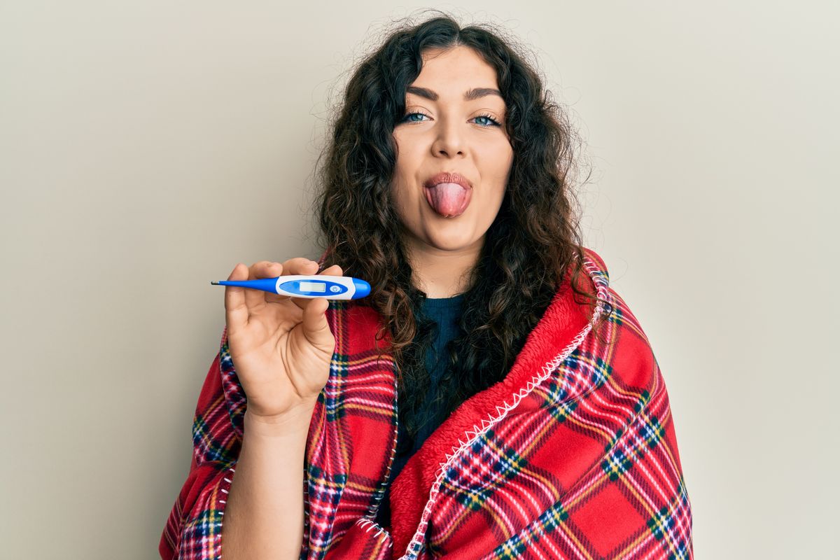 Young brunette woman with curly hair wrapped in a blanket holding thermometer sticking tongue out happy with funny expression. hipochonder