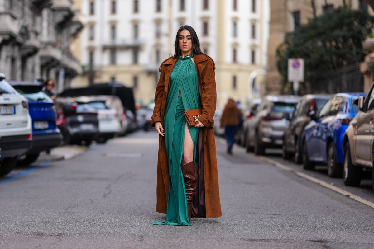 MILAN, ITALY - FEBRUARY 28: A guest wears dark brown suede trench coat, dark green draped halter top with silver detail, dark green high waisted high slit maxi skirt, shiny dark brown knee high slouchy boots leather shoes, dark brown clutch leather bag, outside Sportmax, during the Milan Fashion week Women's Fall/Winter 2025-2026 on February 28, 2025 in Milan, Italy. (Photo by Edward Berthelot/Getty Images)