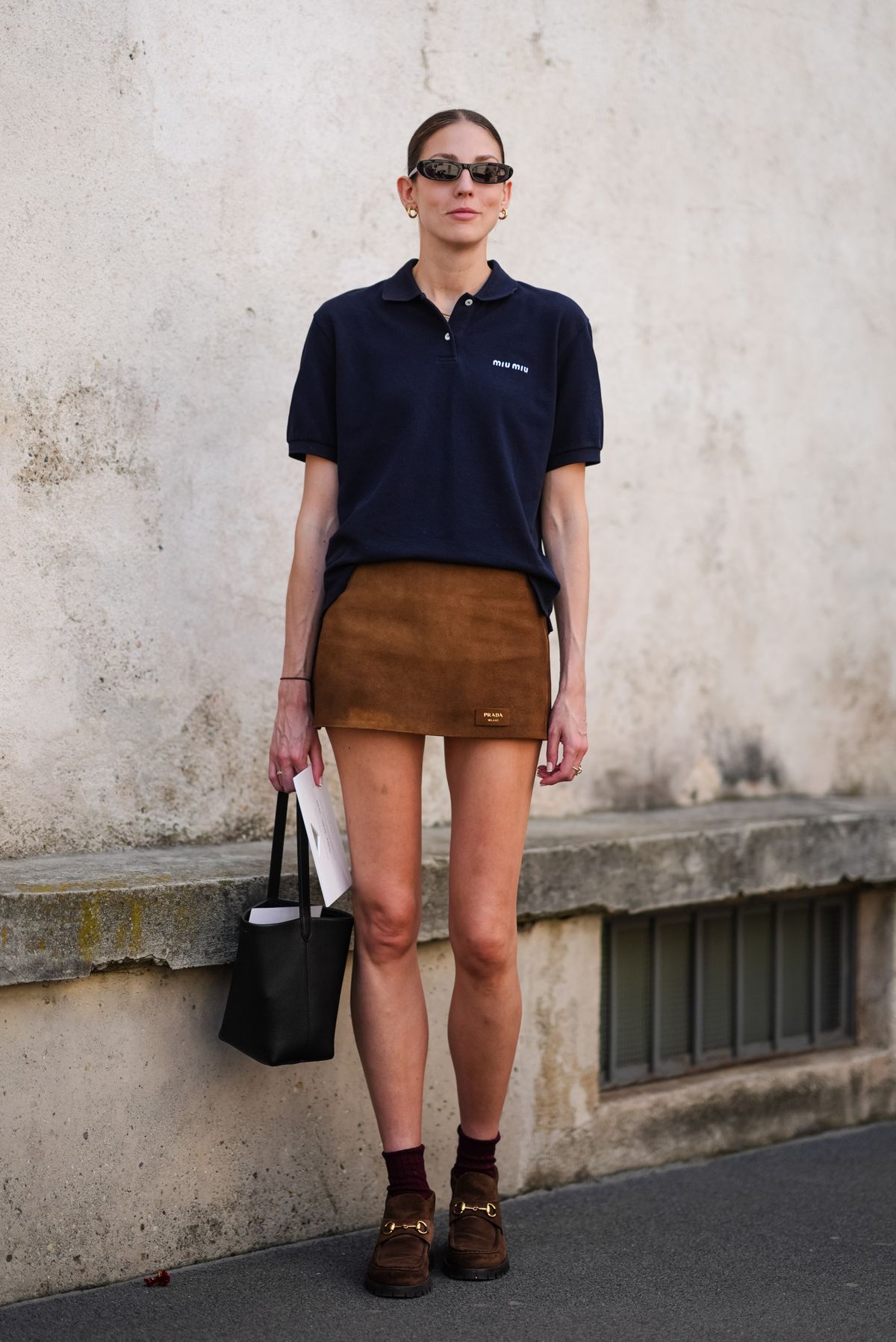 MILAN, ITALY - JUNE 16: A guest wears black sunglasses, gold hoop earrings, black three buttoned short sleeve cotton shirt, brown suede mini skirt, silver rings, black small bag, red socks, suede brown loafers, outside Prada, during the Milan Fashion Week Menswear Spring/Summer 2025 on June 16, 2024 in Milan, Italy. (Photo by Edward Berthelot/Getty Images)