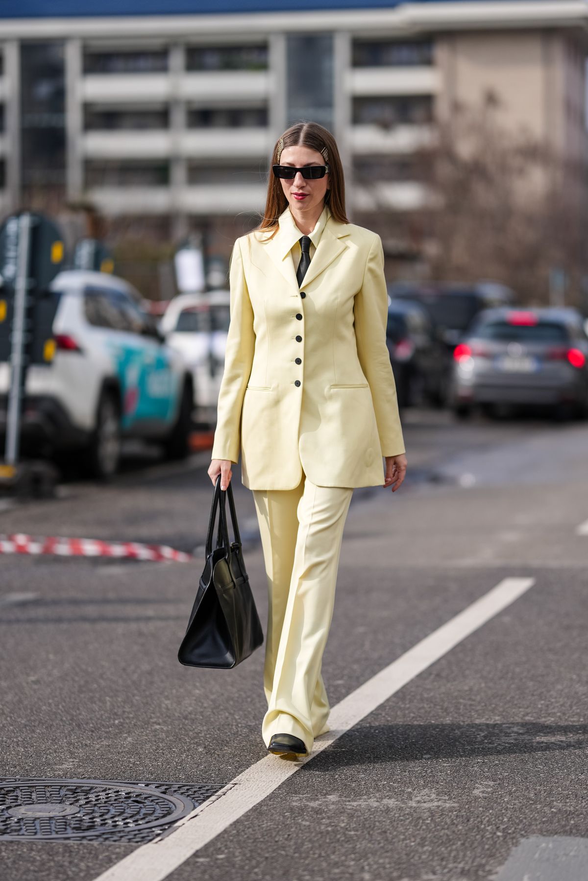 MILAN, ITALY - FEBRUARY 26: A guest wears black sunglasses, silver hairpins, light pale yellow buttoned up long sleeve shirt, black necktie, light pale yellow tailored Jil Sander blazer jacket, light pale yellow flared trouser pants, shiny black leather tote bag, black boots leather shoes, outside Jil Sander, during the Milan Fashion week Women's Fall/Winter 2025-2026 on February 26, 2025 in Milan, Italy. (Photo by Edward Berthelot/Getty Images)