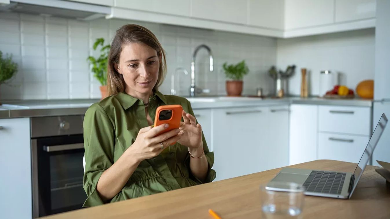 Pleased woman distracted from working on laptop by message sms on mobile phone. Procrastination in remote job from home due to dependence addiction on content on phone realise small amount of dopamine