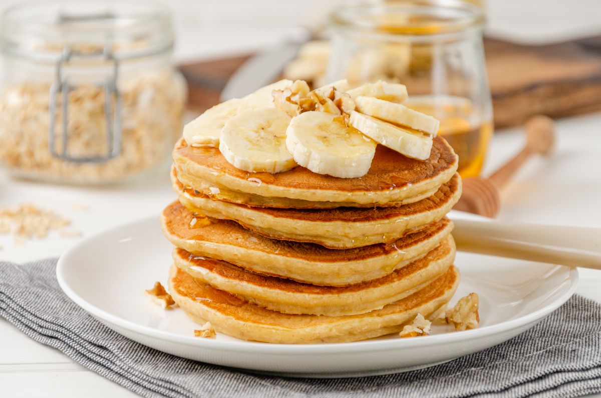 A stack of oatmeal banana pancakes with slices of fresh bananas, walnuts and honey on top with cup of tea on a white wooden background. A healthy breakfast. Copy space