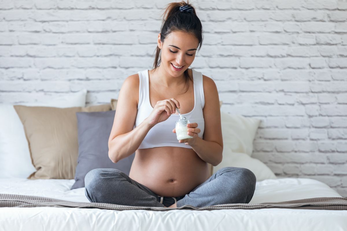 Shot of beautiful young pregnant woman eating yogurt while sitting on the bed at home.