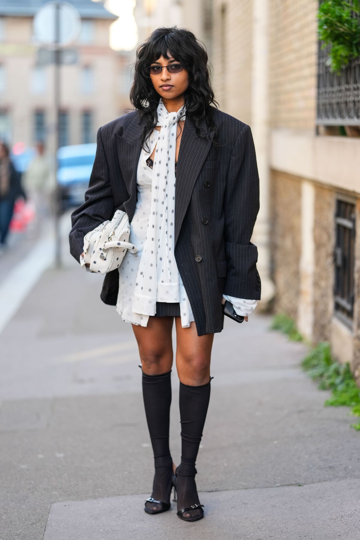 PARIS, FRANCE - MARCH 05: A guest wears black sunglasses, white dark gray polka dotted scarf long sleeve mini dress, black white pinstripe double breasted oversized blazer jacket, white dark gray polka dotted leather bag, black knee high socks, shiny black open toe mules leather shoes, outside Acne Studios, during the Paris Fashion week Women's Fall/Winter 2025-2026 on March 5, 2025 in Paris, France. (Photo by Edward Berthelot/Getty Images)