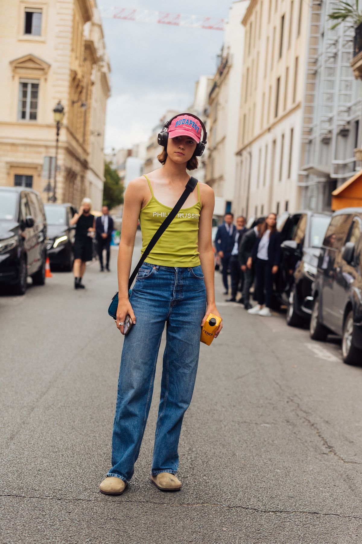 PARIS, FRANCE - JULY 07: Model Dana Smith wears black Marshall headphones, a coral Budapest Original cap, green Philip Russel tank top, blue jeans, tan suede Birkenstock sandals, and carries a yellow Fendi boxed water at the Fendi show at Palais Brongniart  during Couture Fashion Week Fall/Winter 2022 on July 07, 2022 in Paris, France. (Photo by Melodie Jeng/Getty Images)