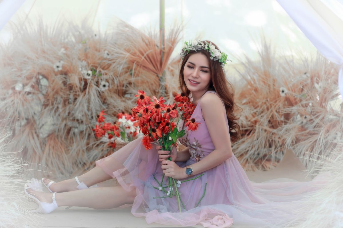 A woman in a soft pink dress, wearing a floral crown, holds a bouquet of bright orange flowers in a peaceful, dreamy atmosphere surrounded by dried plants.