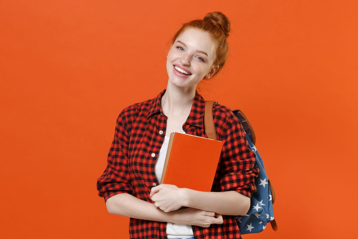 Smiling young teen redhead student girl in casual red checkered shirt backpack posing isolated on orange background. Education in high school university college concept. Mock up copy space. Hold books