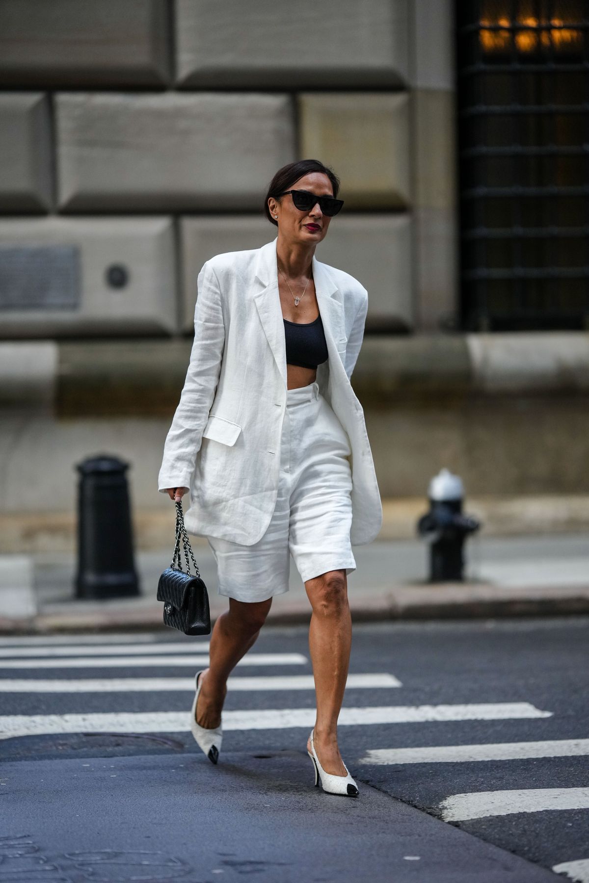 NEW YORK, NEW YORK - SEPTEMBER 13: A guest wears black sunglasses, diamond earrings, a gold chain with white crystal pendant necklace, a black ribbed cropped top, a white oversized linen blazer jacket, high waist white linen shorts, a black shiny leather Timeless handbag from Chanel, white tweed with black shiny varnished leather pointed pumps heels shoes from Chanel , outside Peter Do, during New York Fashion Week, on September 13, 2022 in New York City. (Photo by Edward Berthelot/Getty Images)