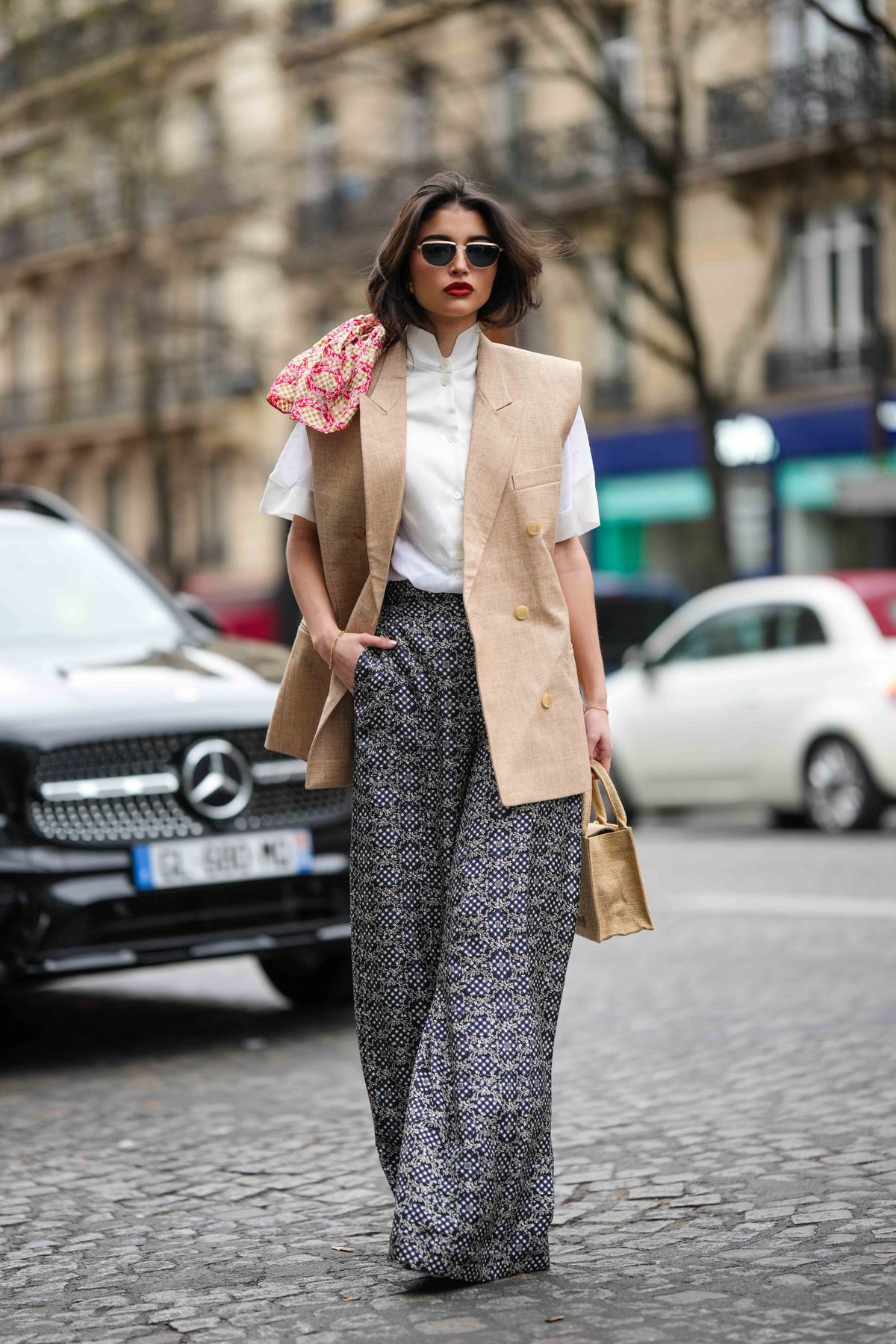 PARIS, FRANCE - MARCH 05: A guest wears black and gold sunglasses, a white high neck buttoned blouse short sleeves shirt, a beige linen sleeveless blazer gilet, a beige wicker handbag, a white and black print pattern large flowing pants, pumps heels shoes , outside EENK, during Paris Fashion Week - Womenswear Fall Winter 2023 2024, on March 05, 2023 in Paris, France. (Photo by Edward Berthelot/Getty Images)