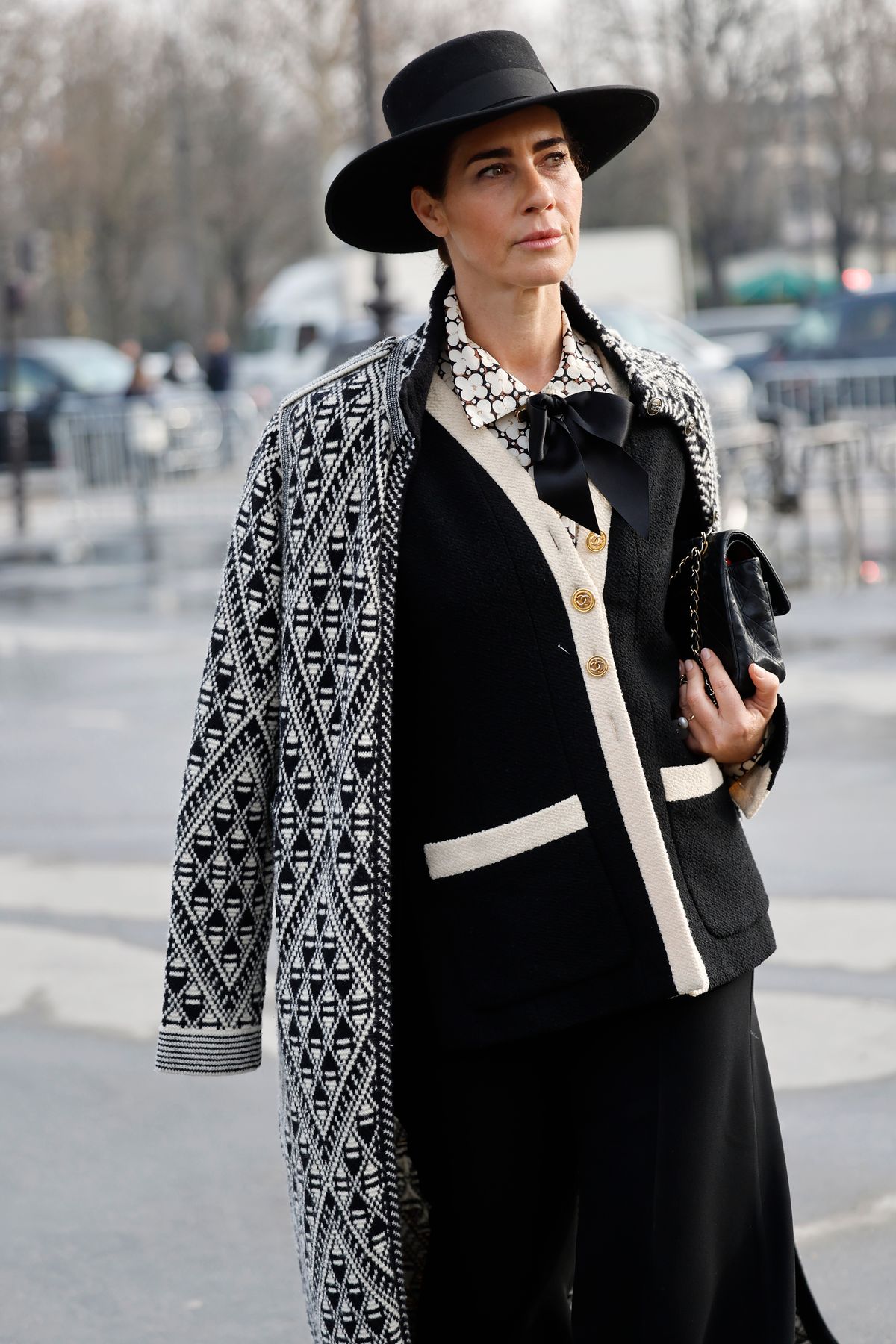 PARIS, FRANCE - MARCH 11: A guest wears black cardigan with cream details, black trousers, black hat, black Chanel bag, black and white printed coat, outside Chanel, during the Womenswear Fall/Winter 2025/2026 as part of Paris Fashion Week on March 11, 2025 in Paris, France. (Photo by Claudio Lavenia/Getty Images)