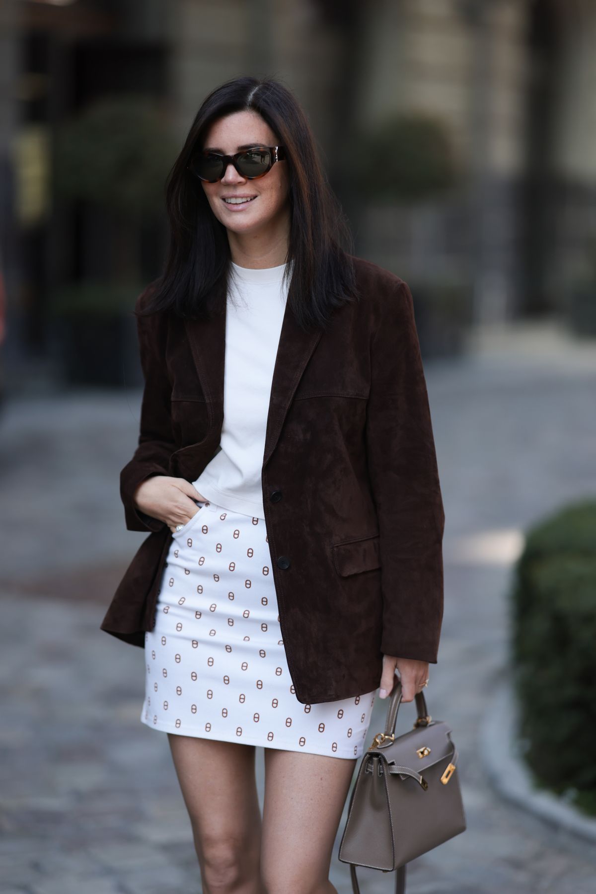 BERLIN, GERMANY - APRIL 02: Leo Eberlin seen wearing Yves Saint Laurent black oversized sunglasses giving off a confident, urban-cool vibe, Leo Mathild diamond jewelry, COS simple, clean white cotton t-shirt, Hermès white denim mini skirt that features a repeating pattern, Zara chocolate brown suede leather blazer, Hermès small, structured taupe leather Kelly handbag with gold hardware, on April 02, 2025 in Berlin, Germany. (Photo by Jeremy Moeller/Getty Images)