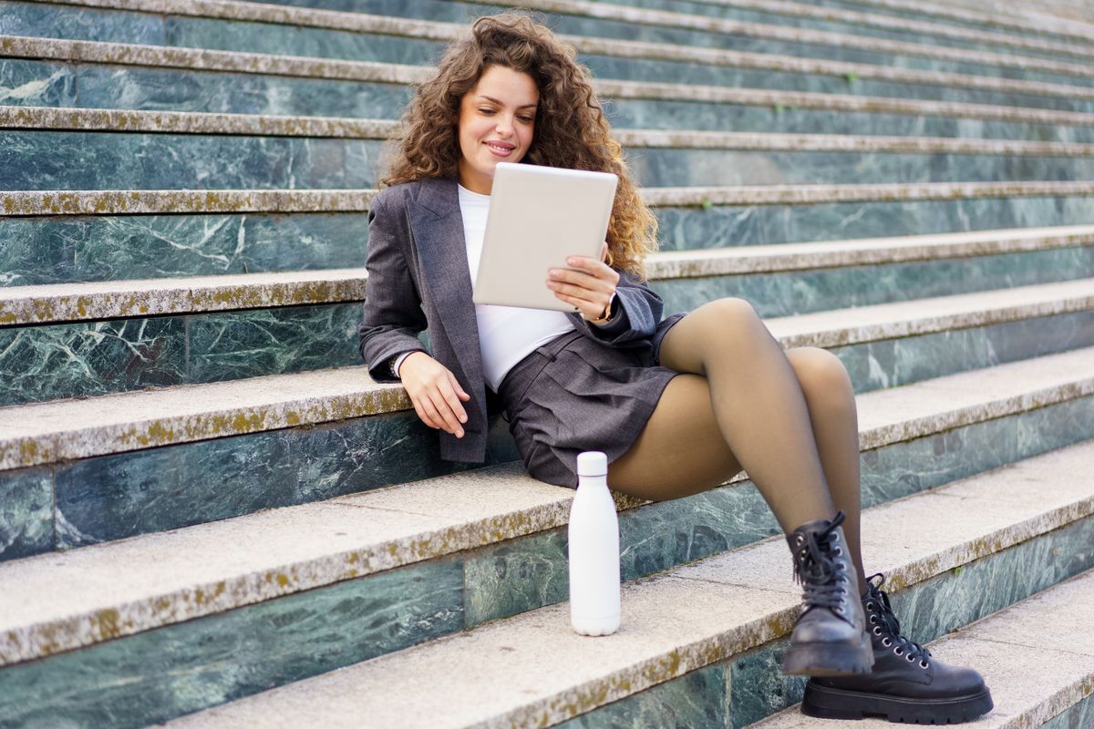 A young woman sits on outdoor stairs, engrossed in a captivating book, dressed stylishly, holding a trendy water bottle, embodying a fashionable casual lifestyle many aspire to emulate