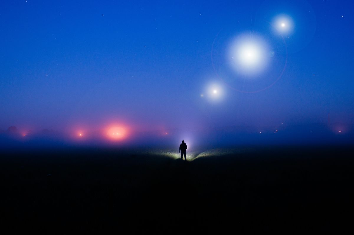 A silhouette of a figure, standing in the countryside on a foggy night, with glowing lights of UFO's in the sky