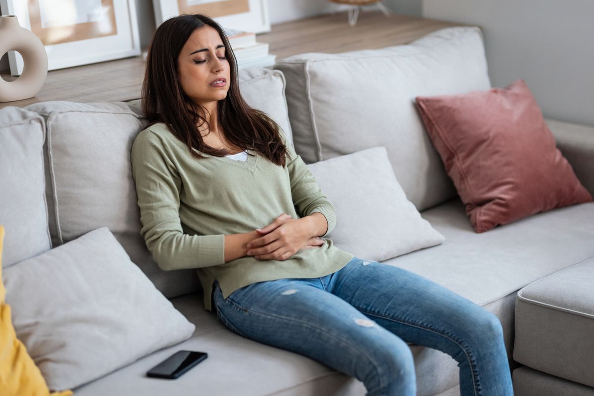 Shot of unhealthy young woman with stomachache leaning on the couch in the living room at home.
