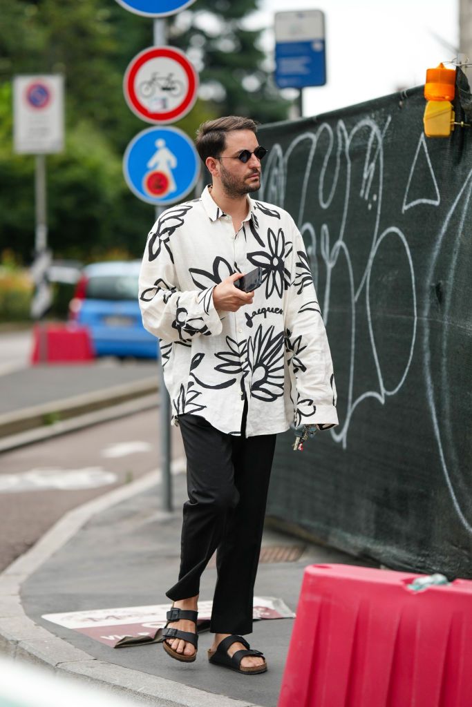 MILAN, ITALY - JUNE 18: A guest wears black circle sunglasses, a white with embroidered large flower pattern shirt from Jacquemus, black suit pants, black shiny leather belted sandals , outside JW Anderson, during the Milan Fashion Week - Menswear Spring/Summer 2024 on June 18, 2023 in Milan, Italy. (Photo by Edward Berthelot/Getty Images)