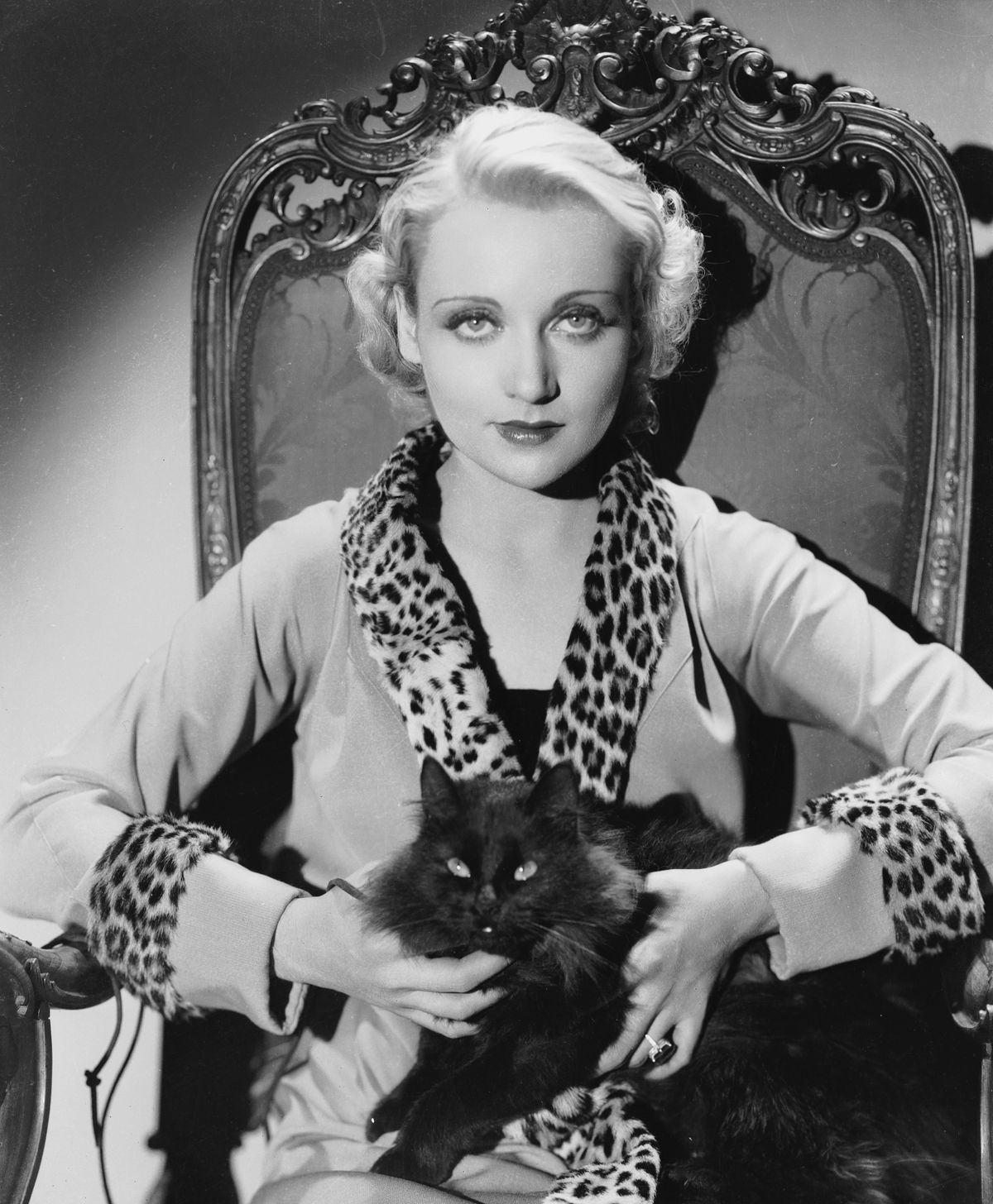 circa 1930:  American film actress Carole Lombard  (1908 - 1942) wearing a jacket with a leopard skin collar and cuffs, and holding a black cat on her lap.  (Photo via John Kobal Foundation/Getty Images)