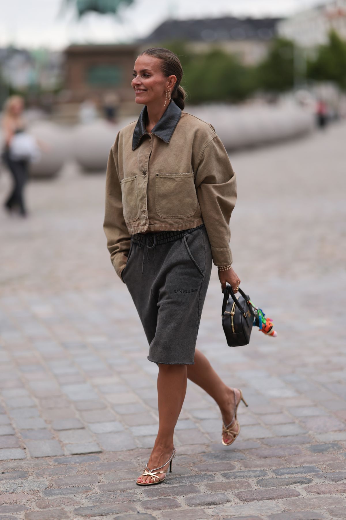 COPENHAGEN, DENMARK - AUGUST 08: Janka Polliani is seen wearing one hanging gold earring with rhinestones and the matching bracelet from Dulong Fine Jewelry, a cropped brown jeans jacket with dark brown leather collar and large pockets from Prada, washed-out grey knee-length wide shorts from H2OFagerhol, two gold rings, a rectangular black leather bag from Miu Miu with several multicolored bag charms, and shiny golden heeled strap sandals before the Rotate Show during day four of the Copenhagen Fashion Week (CPHFW) SS25 on August 08, 2024 in Copenhagen, Denmark. (Photo by Jeremy Moeller/Getty Images)