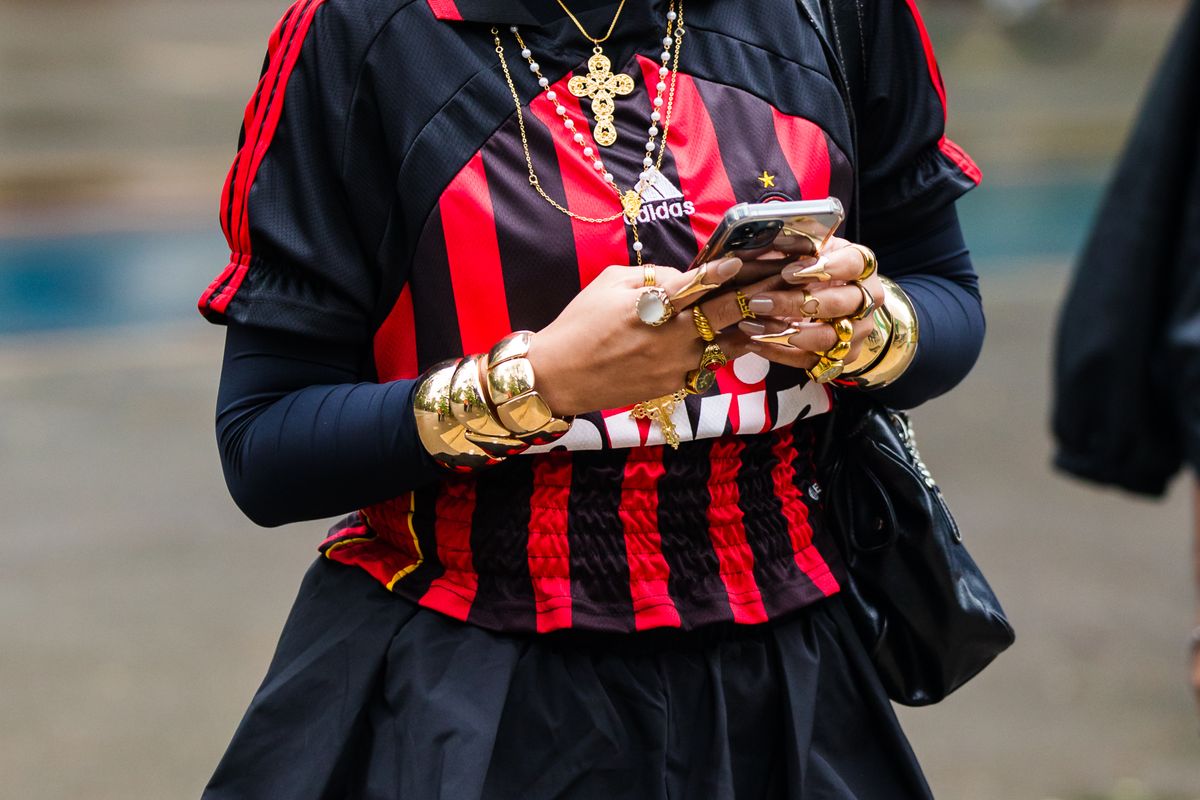 SAO PAULO, BRAZIL - OCTOBER 20: A guest wears football jersey, red and black vertical stripes, layered long-sleeve top, gold statement bracelets, stacked rings, gold cross necklace, pearl necklace, pleated black skirt, oversized black handbag, bold gold accessories, intricate nail design, sporty and elegant elements, casual yet accessorized look at Sao Paulo Fashion Week 2024 N58 - Street Style at Parque Ibirapuera on October 20, 2024 in Sao Paulo, Brazil. (Photo by Mauricio Santana/Getty Images)