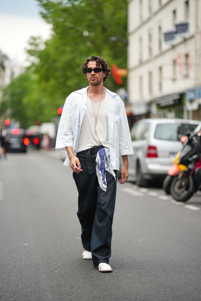 PARIS, FRANCE - JUNE 21: Yanis Serbout wears black sunglasses, a silver chain and long pendant necklace, a white t-shirt, a white gray cotton long short sleeves shirt, navy blue denim large pants, a white and navy blue print pattern silk scarf as a belt, white shiny leather strappy sandals , outside Bluemarble, during the Menswear Spring/Summer 2024 as part of  Paris Fashion Week on June 21, 2023 in Paris, France. (Photo by Edward Berthelot/Getty Images)
