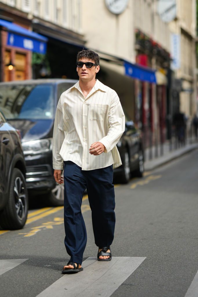 PARIS, FRANCE - JUNE 22: A guest wears black sunglasses, a white latte with small black striped print pattern oversized shirt, navy blue suit pants, black shiny leather strappy sandals, outside the Walter Van Beirendonck show, during Paris Fashion Week - Menswear Spring/Summer 2023, on June 22, 2022 in Paris, France. (Photo by Edward Berthelot/Getty Images)