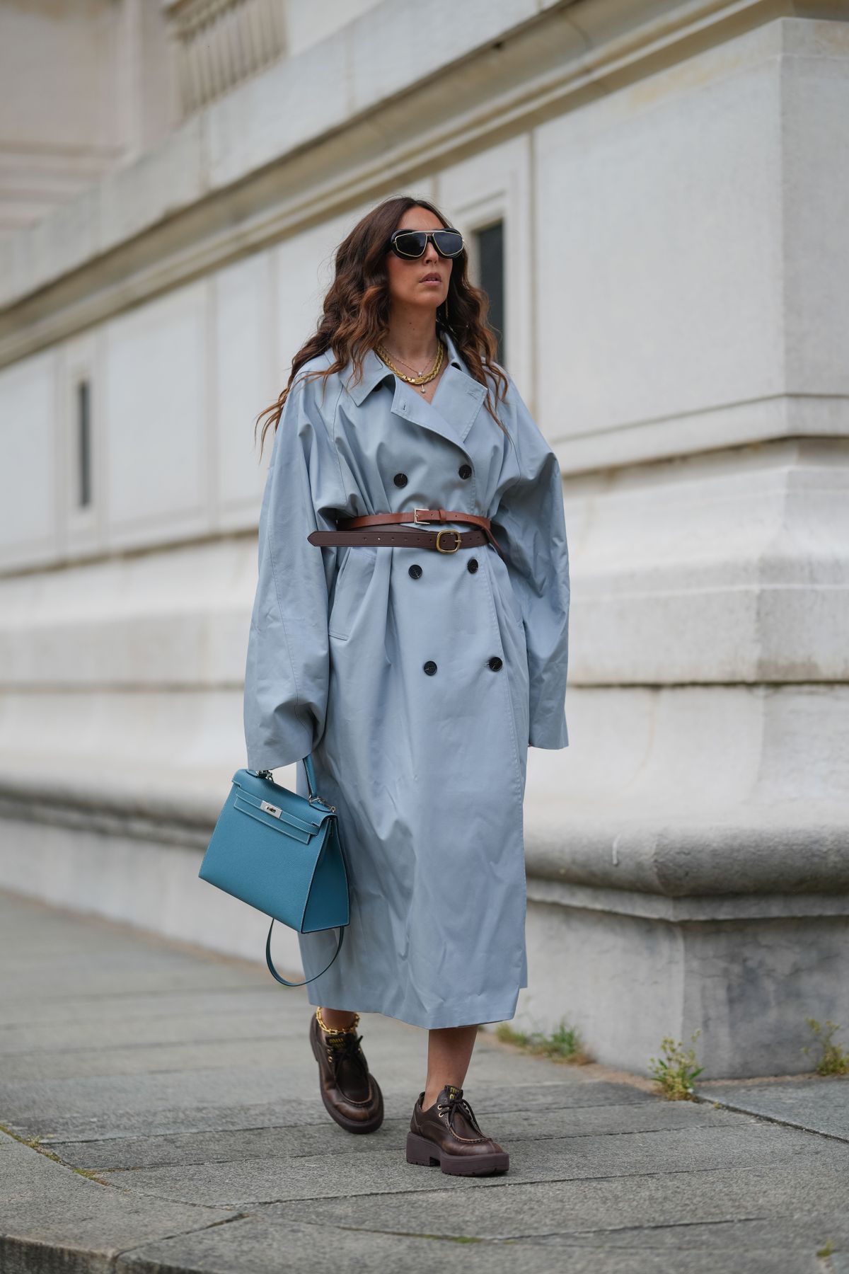 PARIS, FRANCE - APRIL 25: Gabriella Berdugo wears loose, wavy, long brown hair with a middle part, a light blue oversized double-breasted trench coat by Eleh cinched at the waist with a brown leather belt by Ralph Lauren, dark brown chunky lace-up shoes by Miu Miu, black oversized sunglasses by Bottega Veneta with gold detailing, a gold chain anklet by Brink & Eliza, and a turquoise leather top-handle bag by Hermes, captured in a full body shot, during a street style fashion photo session, on April 25, 2025 in Paris, France. (Photo by Edward Berthelot/Getty Images)