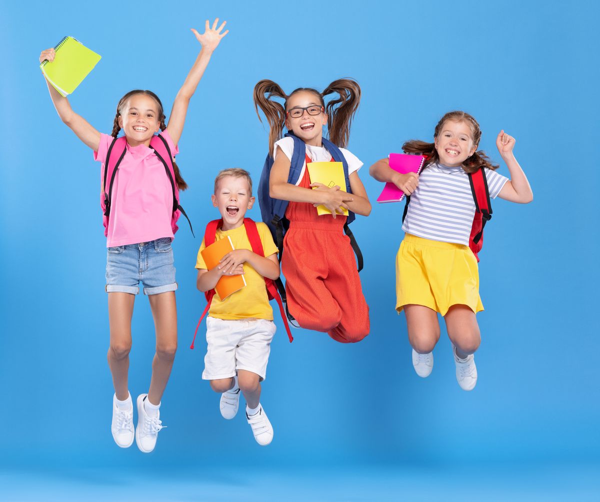 Group of funny excited classmates in casual clothes with backpacks and copybooks having fun and jumping on blue background