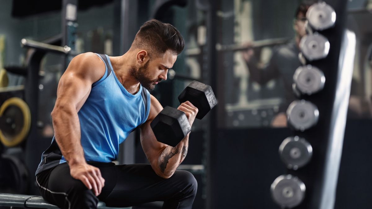 Bodybuilder pumping his biceps with dumbbell in gym, empty space