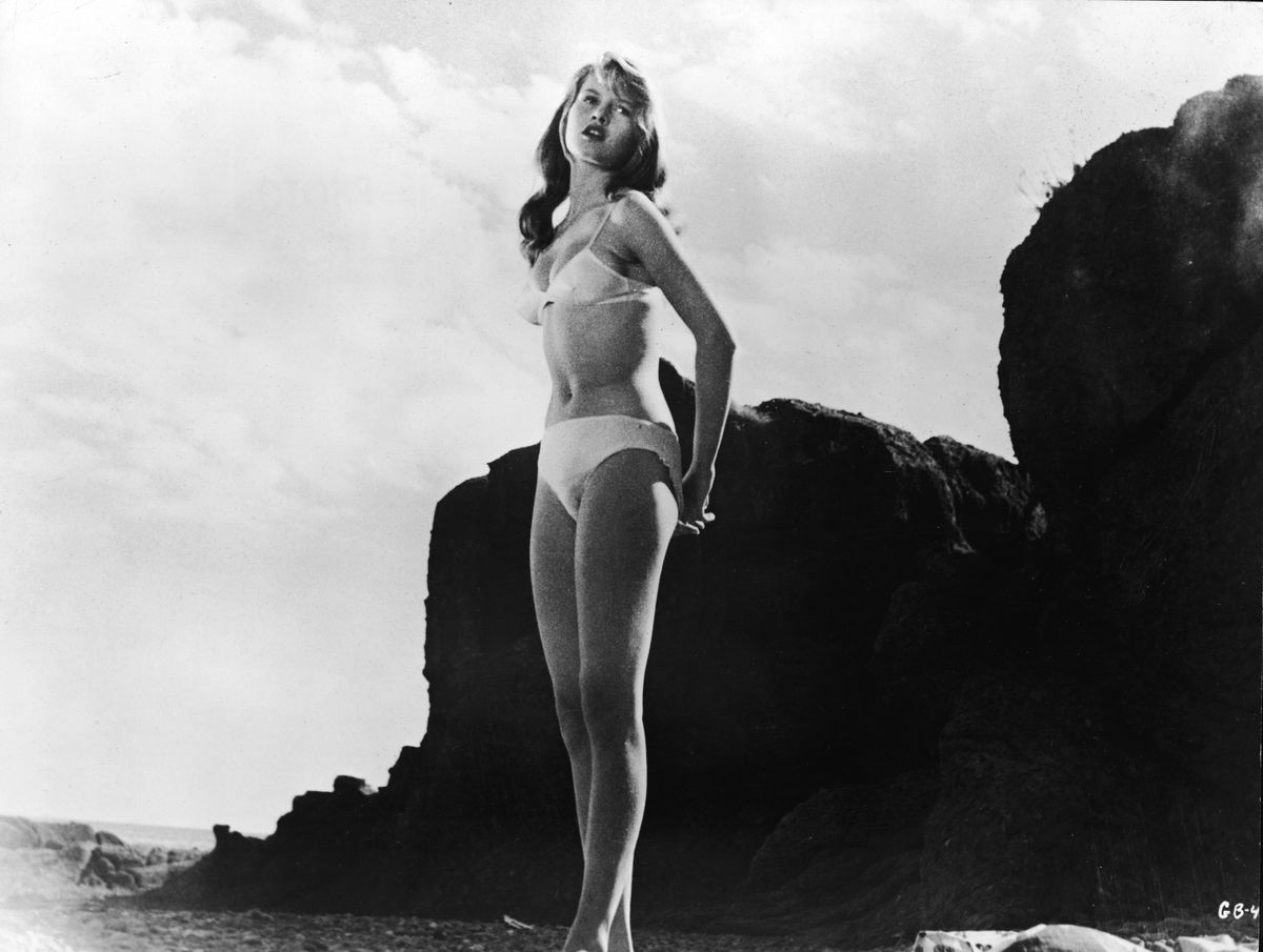 French-born actor Brigitte Bardot wears a white bikini and stands on a rocky beach in a still from the film, 'The Girl in the Bikini',  directed by Willy Rozier, 1958. (Photo by Atlantis Films/Pictorial Parade/Courtesy of Getty Images)