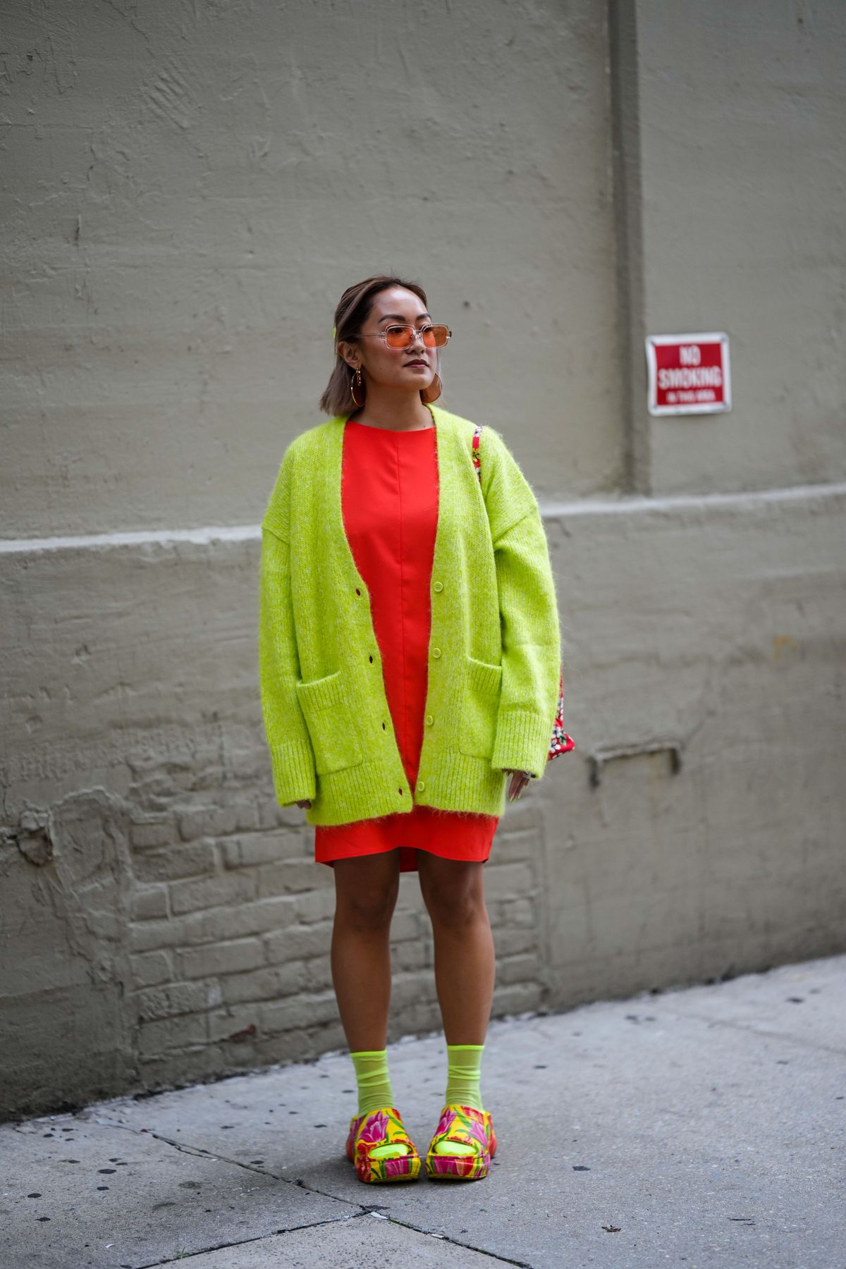 NEW YORK, NEW YORK - SEPTEMBER 13: A guest wears orange sunglasses, gold pendant Fendi earrings, gold earrings, a neon orange short dress, a neon yellow oversized wool cardigan, neon yellow socks, a red with white flower print pattern shoulder bag, yellow with red and pink flower painted pattern open toe cap / plastic mules , outside COS, during New York Fashion Week, on September 13, 2022 in New York City. (Photo by Edward Berthelot/Getty Images)