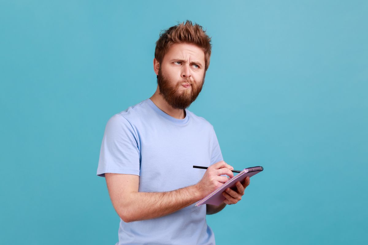 Portrait of bearded man making notes in paper notebook, writing business idea, future plans, looking away with thoughtful expression. Indoor studio shot isolated on blue background.