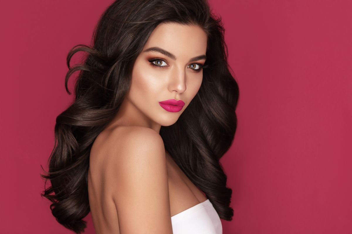 Brunette woman with voluminous curls and bold pink lipstick poses against a pink background