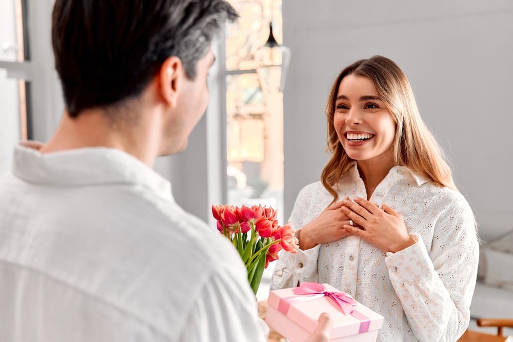 trend A man is congratulating his wife by giving her a bouquet of flowers and a gift. His excited and happy wife is rejoicing. The loving couple is at home in a bright apartment.