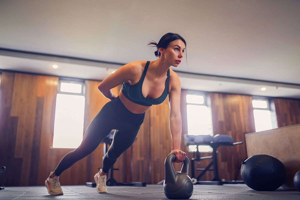 young motivated girl doing plank exercise using kettlebells with one hand at gym, full length photo, copy space.