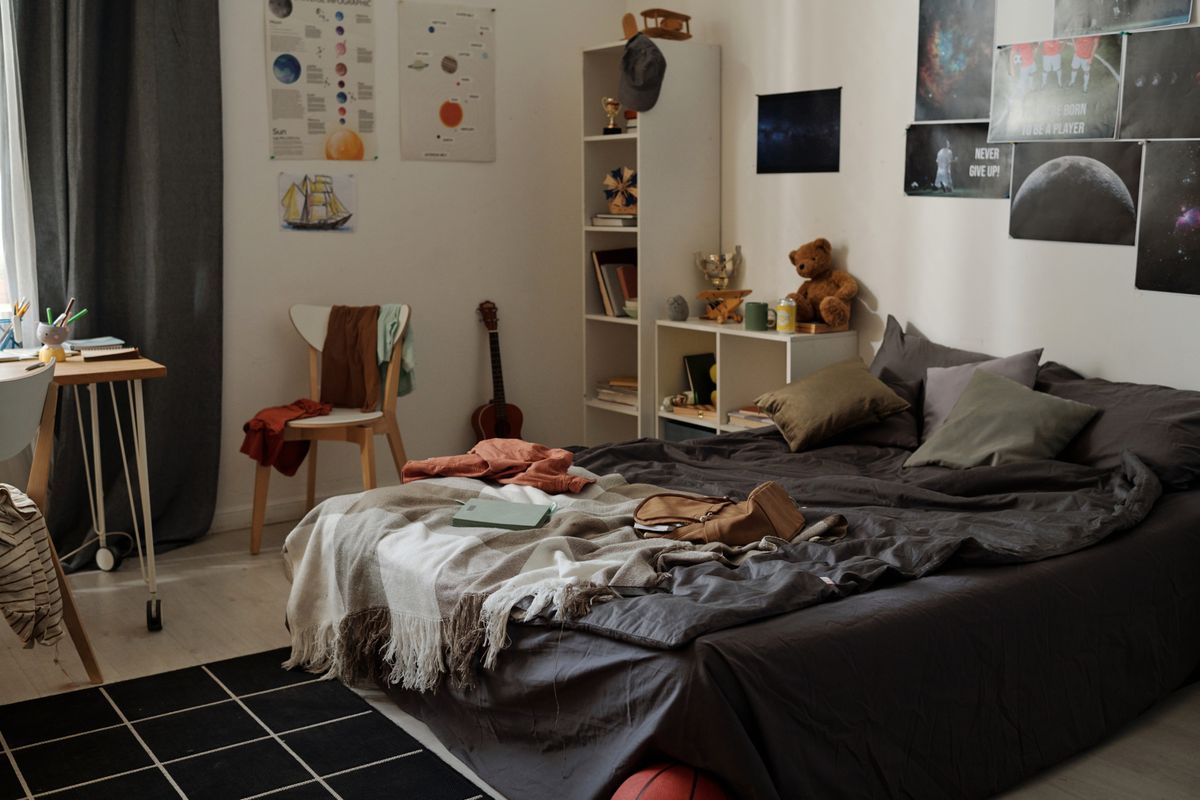 Messy double bed with cushions, blanket, plaid and backpack standing by wall with posters and shelves with various stuff in bedroom