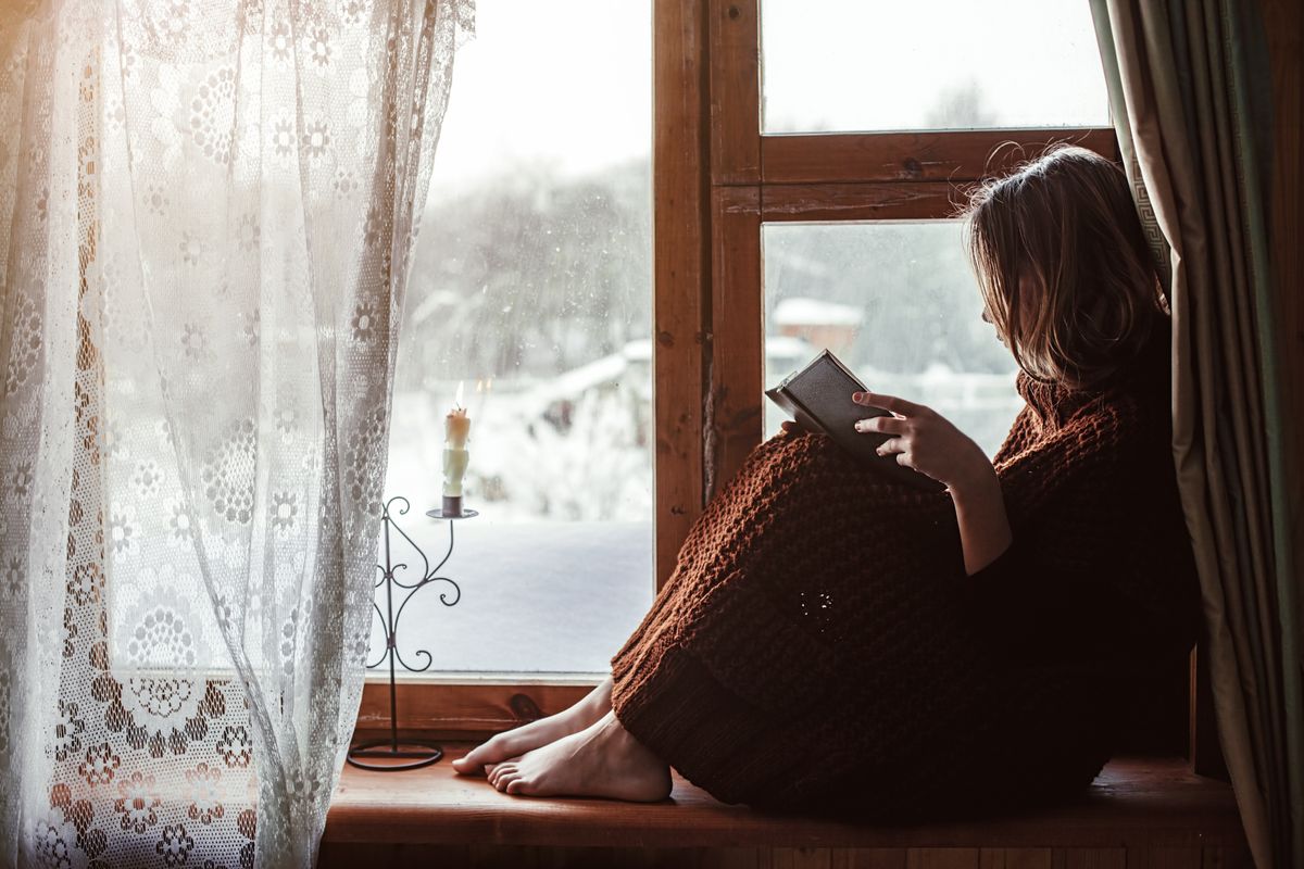 Pre teen child in warm woolen sweater seating on window sill and reading a book. Winter weekends in old log house. Cold snowy weather. Cozy homely concept.