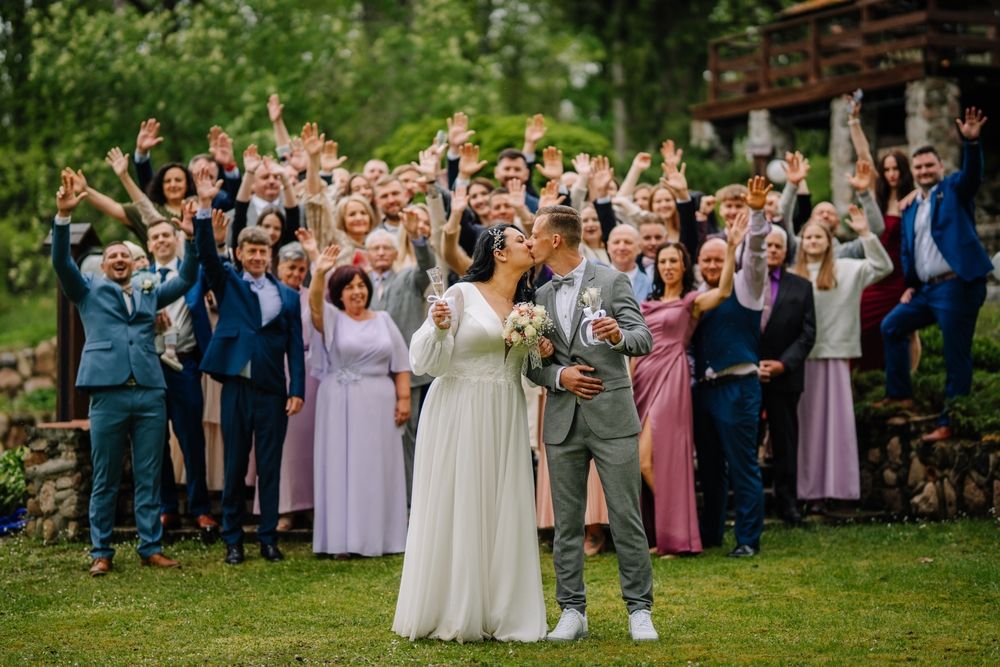 menyasszony Valmiera, Latvia - May 23, 2025 - Bride and groom kiss while holding champagne and bouquet, surrounded by cheering guests in a lively outdoor wedding group photo.