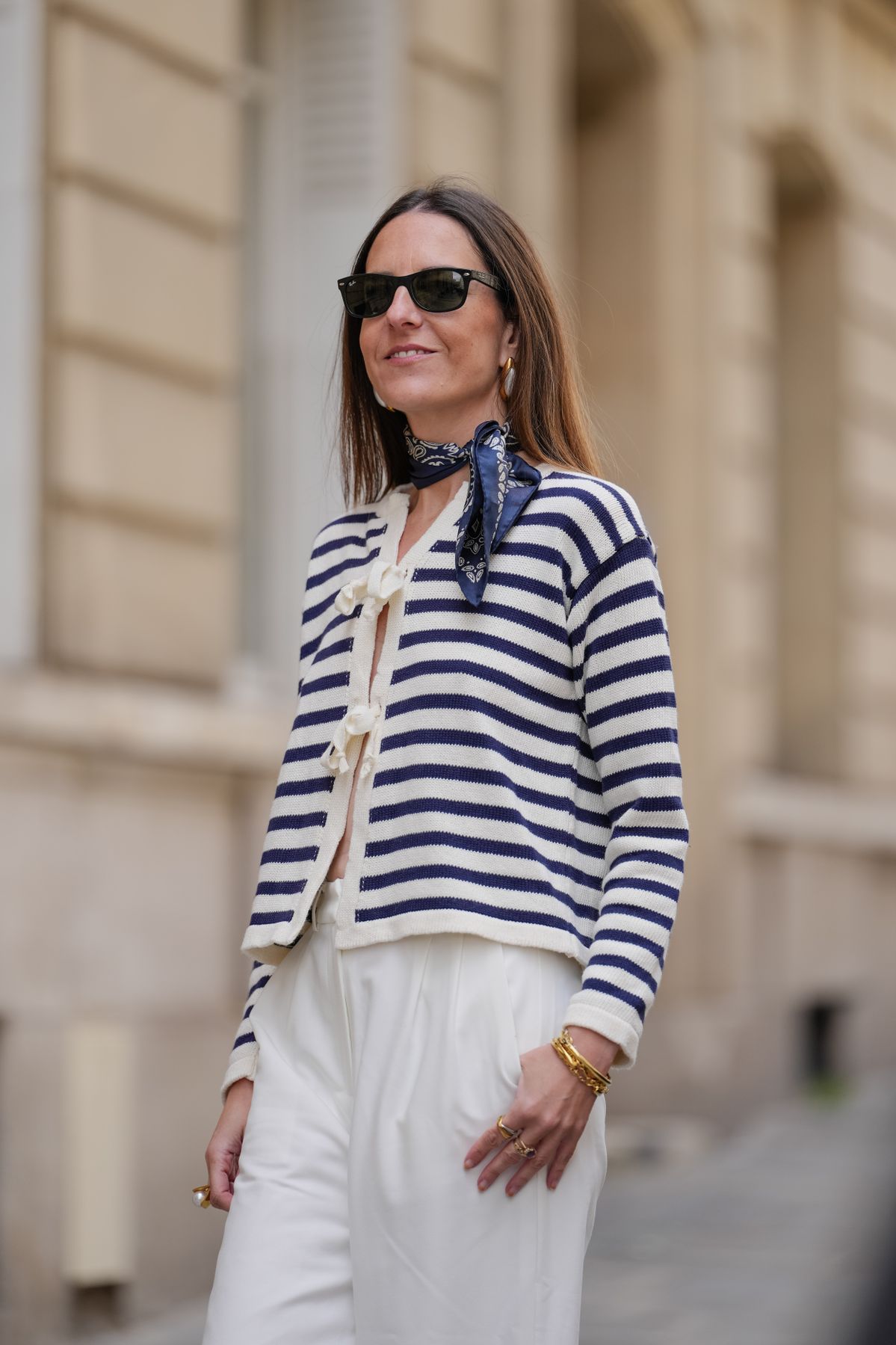 PARIS, FRANCE - MAY 07: Alba Garavito Torre wears long, straight brown hair with a middle part, dark pink lipstick and defined brows. Black square sunglasses by Ray-Ban are styled with a navy printed scarf tied around the neck and gold hoop earrings. A navy and beige striped cardigan with wool ties by Frambua is paired with white wide-leg pants. This is a mid shot, during a street style fashion photo session, on May 07, 2025 in Paris, France. (Photo by Edward Berthelot/Getty Images)