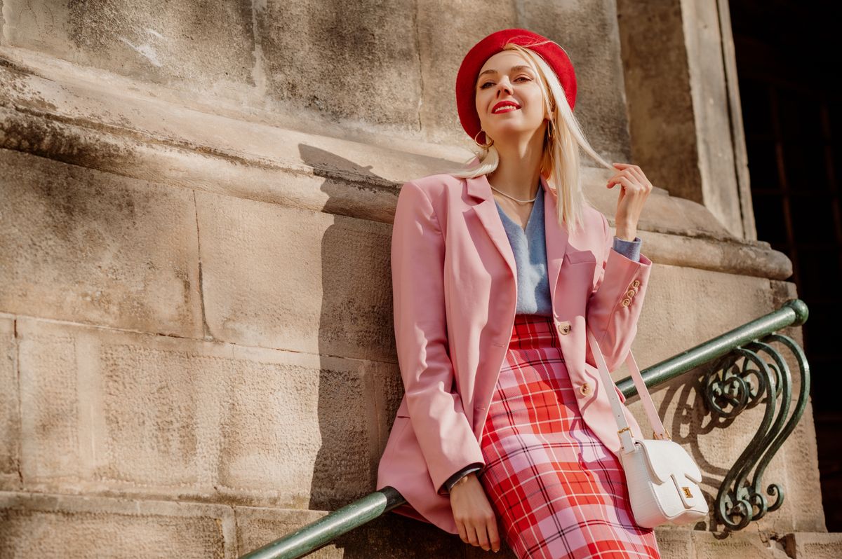 Fashionable happy smiling blonde woman wearing stylish red beret, classic pink suit blazer, checkered skirt, holding trendy white bag, posing in street of European city. Copy, empty space for text