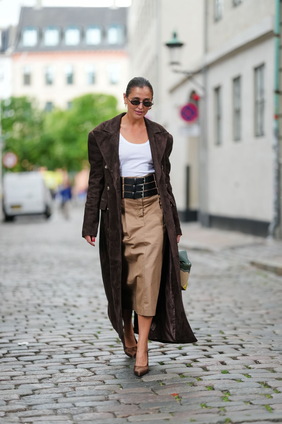 COPENHAGEN, DENMARK - AUGUST 04: Darja Barannik wears a long brown suede coat with wide lapels and double-breasted gold buttons. Underneath, a white scoop-neck tank-top is visible. A high-waisted beige skirt extends to mid-calf. A wide black belt with multiple buckles cinches the waist. Accessories include small oval sunglasses and gold hoop earrings. The green handbag features a geometric design with beige and yellow accents and a braided handle from Loewe. Brown pointed-toe heels complete the ensemble, outside Freya Dalsjo, during Copenhagen Fashion Week, on August 04, 2025 in Copenhagen, Denmark (Photo by Edward Berthelot/Getty Images)